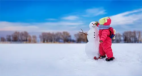 Petite fille en combinaison de ski construisant un bonhomme de neige, symbole des vacances d’hiver en famille