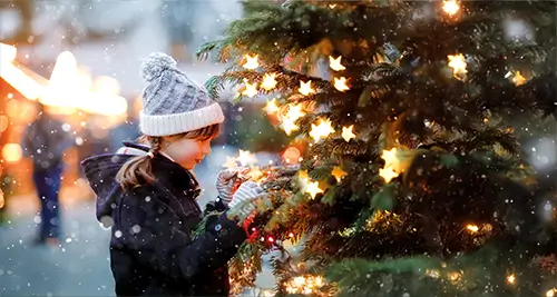 Petite fille accrochant une boule de Noël à un sapin illuminé en devant un des villages de vacances en France VTF