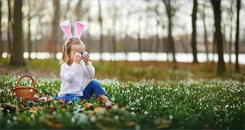 Petite fille avec des oreilles de lapin jouant avec des œufs de Pâques dans l’herbe, symbole des villages de vacances France au printemps